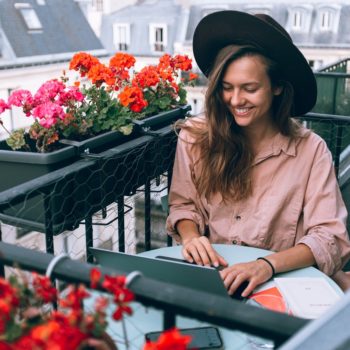 Woman in hat sitting outside smiling at her laptop