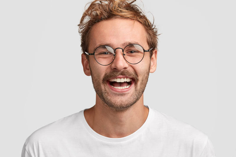 Close up shot of happy stylish male in round glasses, has positive smile on face, glad to recieve salary, going to spend money on new purchases, isolated over white background. People and lifestyle